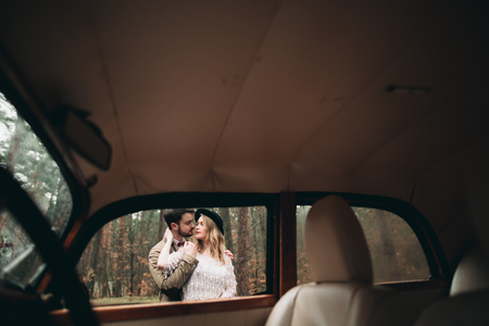 Gorgeous newlywed bride and groom posing in pine forest near retro car in their wedding day.の写真素材