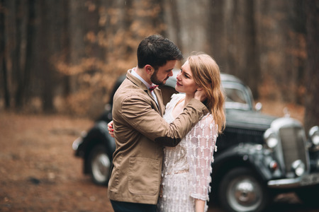 Gorgeous newlywed bride and groom posing in pine forest near retro car in their wedding day.の写真素材