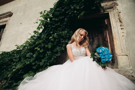 Beautiful young blond bride with bridal bouquet sitting on the stairs under gorgeous plants.の写真素材