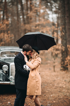 Romantic fairytale wedding couple, bride and groom, kissing and embracing in pine forest near retro car.の写真素材