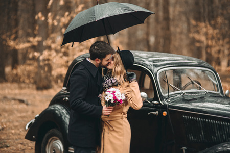 Romantic fairytale wedding couple, bride and groom, kissing and embracing in pine forest near retro car.の写真素材