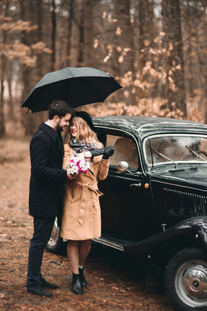 Romantic fairytale wedding couple, bride and groom, kissing and embracing in pine forest near retro car.の写真素材