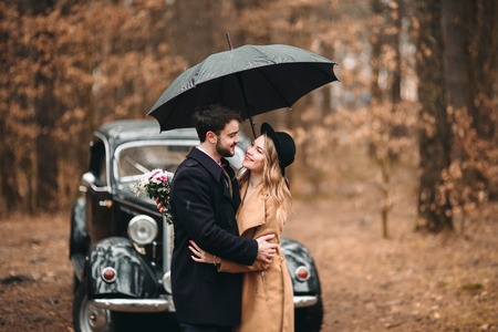 Romantic fairytale wedding couple, bride and groom, kissing and embracing in pine forest near retro car.の写真素材