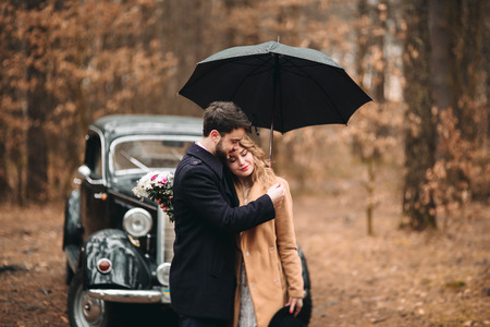 Romantic fairytale wedding couple, bride and groom, kissing and embracing in pine forest near retro car.の写真素材