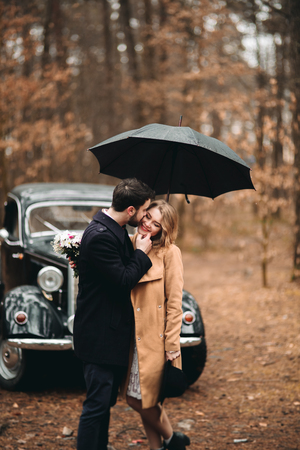 Romantic fairytale wedding couple, bride and groom, kissing and embracing in pine forest near retro car.の写真素材