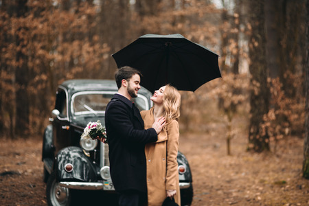 Romantic fairytale wedding couple, bride and groom, kissing and embracing in pine forest near retro car.の写真素材