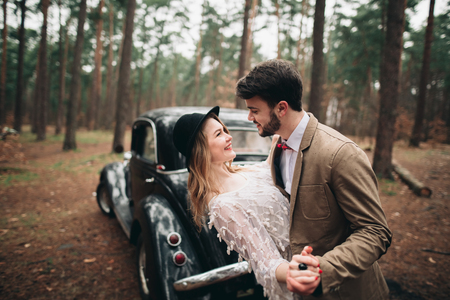 Romantic fairytale wedding couple, bride and groom, kissing and embracing in pine forest near retro car.の写真素材