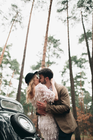 Romantic fairytale wedding couple, bride and groom, kissing and embracing in pine forest near retro car.の写真素材