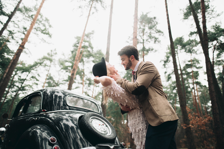 Romantic fairytale wedding couple, bride and groom, kissing and embracing in pine forest near retro car.の写真素材