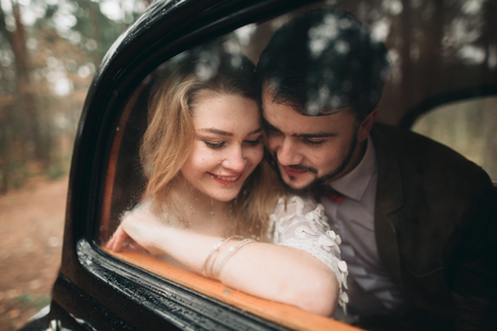 Romantic fairytale wedding couple, bride and groom, kissing and embracing in pine forest near retro car.の写真素材