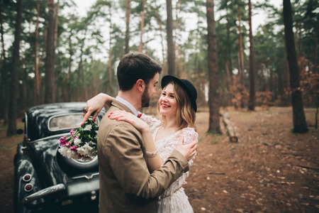 Romantic fairytale wedding couple, bride and groom, kissing and embracing in pine forest near retro car.の写真素材