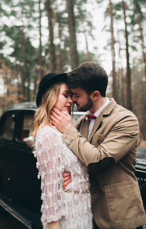 Romantic fairytale wedding couple, bride and groom, kissing and embracing in pine forest near retro car.の写真素材