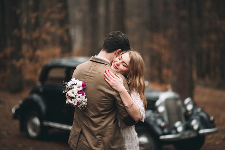 Romantic fairytale wedding couple, bride and groom, kissing and embracing in pine forest near retro car.の写真素材