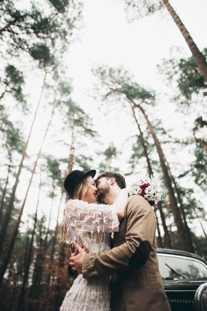 Romantic fairytale wedding couple, bride and groom, kissing and embracing in pine forest near retro car.の写真素材