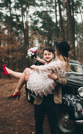 Romantic fairytale wedding couple, bride and groom, kissing and embracing in pine forest near retro car.の写真素材
