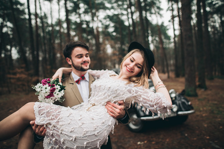 Romantic fairytale wedding couple, bride and groom, kissing and embracing in pine forest near retro car.の写真素材