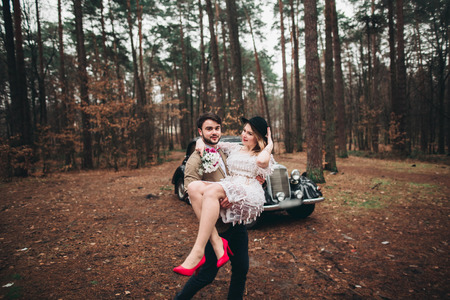 Romantic fairytale wedding couple, bride and groom, kissing and embracing in pine forest near retro car.の写真素材