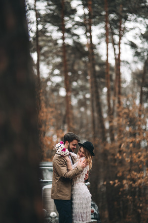 Romantic fairytale wedding couple, bride and groom, kissing and embracing in pine forest near retro car.の写真素材