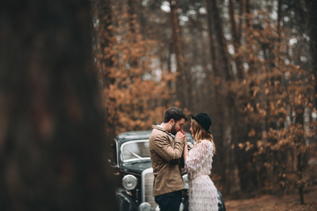 Romantic fairytale wedding couple, bride and groom, kissing and embracing in pine forest near retro car.の写真素材