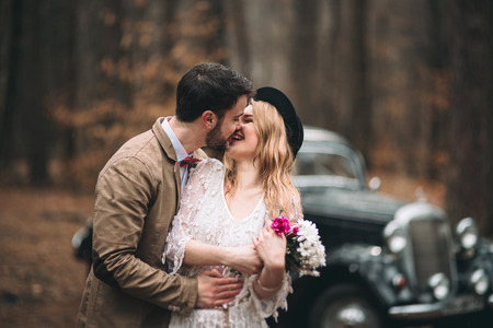 Romantic fairytale wedding couple, bride and groom, kissing and embracing in pine forest near retro car.の写真素材