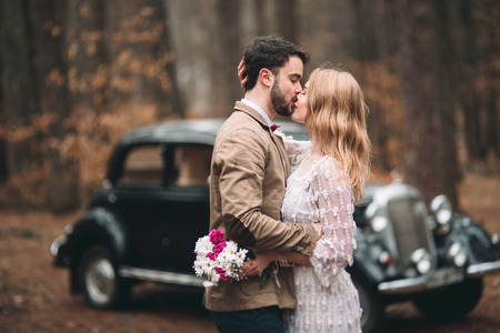 Romantic fairytale wedding couple, bride and groom, kissing and embracing in pine forest near retro car.の写真素材