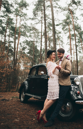Romantic fairytale wedding couple, bride and groom, kissing and embracing in pine forest near retro car.の写真素材
