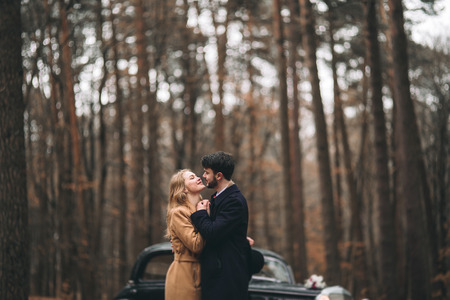 Romantic fairytale wedding couple, bride and groom, kissing and embracing in pine forest near retro car.の写真素材
