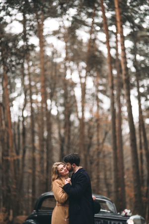 Romantic fairytale wedding couple, bride and groom, kissing and embracing in pine forest near retro car.の写真素材