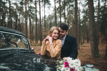 Romantic fairytale wedding couple, bride and groom, kissing and embracing in pine forest near retro car.の写真素材