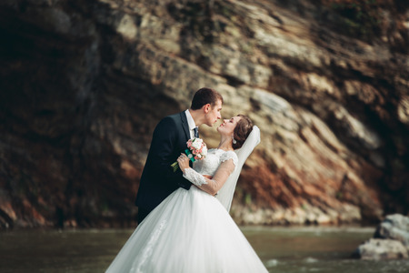 Handsome romantic groom and beautiful bride kissing near river in scenic mountains.の写真素材