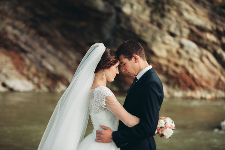 Handsome romantic groom and beautiful bride posing near river in scenic mountains.の写真素材