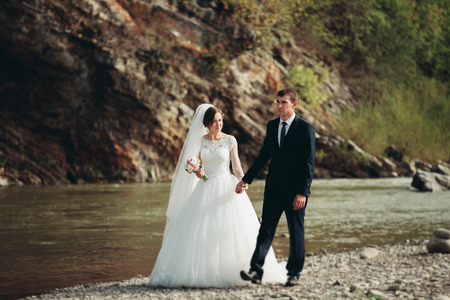 Handsome romantic groom and beautiful bride posing near river in scenic mountains.の写真素材