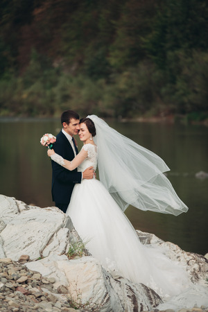 Handsome romantic groom and beautiful bride posing near river in scenic mountains.の写真素材