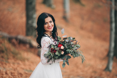 Gorgeous brunette bride in elegant dress holding bouquet and posing near forest and lake.の写真素材