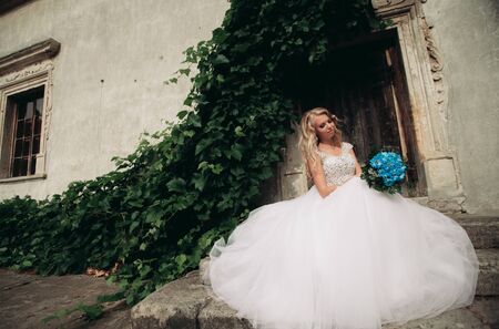 Beautiful young blond bride with bridal bouquet sitting on the stairs background gorgeous plants.の写真素材