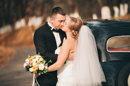 Stylish wedding couple, bride and groom kissing and hugging near retro car in autumn.の写真素材