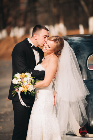 Stylish wedding couple, bride and groom kissing and hugging near retro car in autumn.の写真素材