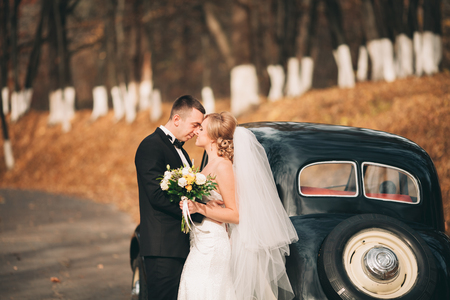 Stylish wedding couple, bride and groom kissing and hugging near retro car in autumn.の写真素材
