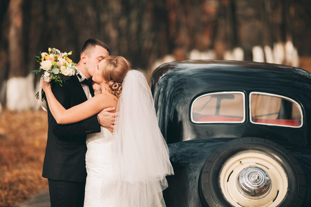 Stylish wedding couple, bride and groom kissing and hugging near retro car in autumn.の写真素材