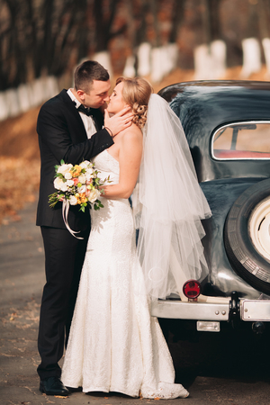 Stylish wedding couple, bride and groom kissing and hugging near retro car in autumn.の写真素材