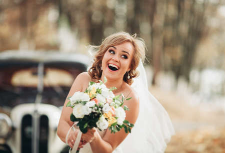Beautiful happy bride with bouquet near retro car in autumn.の写真素材