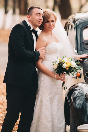 Stylish wedding couple, bride and groom hugging near retro car in autumn.の写真素材