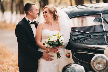 Stylish wedding couple, bride and groom kissing and hugging near retro car in autumn.の写真素材