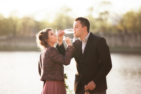 Wedding couple, groom and dress posing with wine glasses near river with a glass at sunset.の写真素材