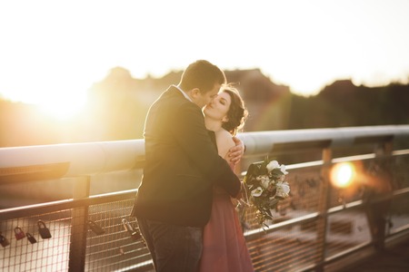 Stylish loving wedding couple, groom, bride with pink dress kissing and hugging on a bridge at...