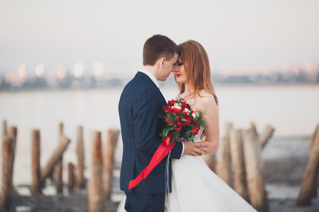 Wedding couple, groom, bride with bouquet posing near sea on sunset.の写真素材