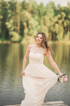 Gorgeous bride in elegant dress holding bouquet posing near forest and lake.の写真素材