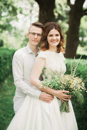 Stylish couple of happy stylish newlyweds walking in the park on their wedding day with bouquet.の写真素材