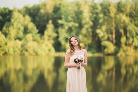 Gorgeous bride in elegant dress holding bouquet posing near forest and lake.の写真素材