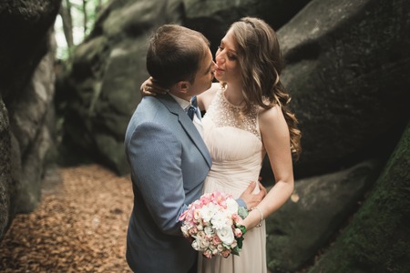Gorgeous wedding couple kissing and hugging in forest with big rocks.の写真素材